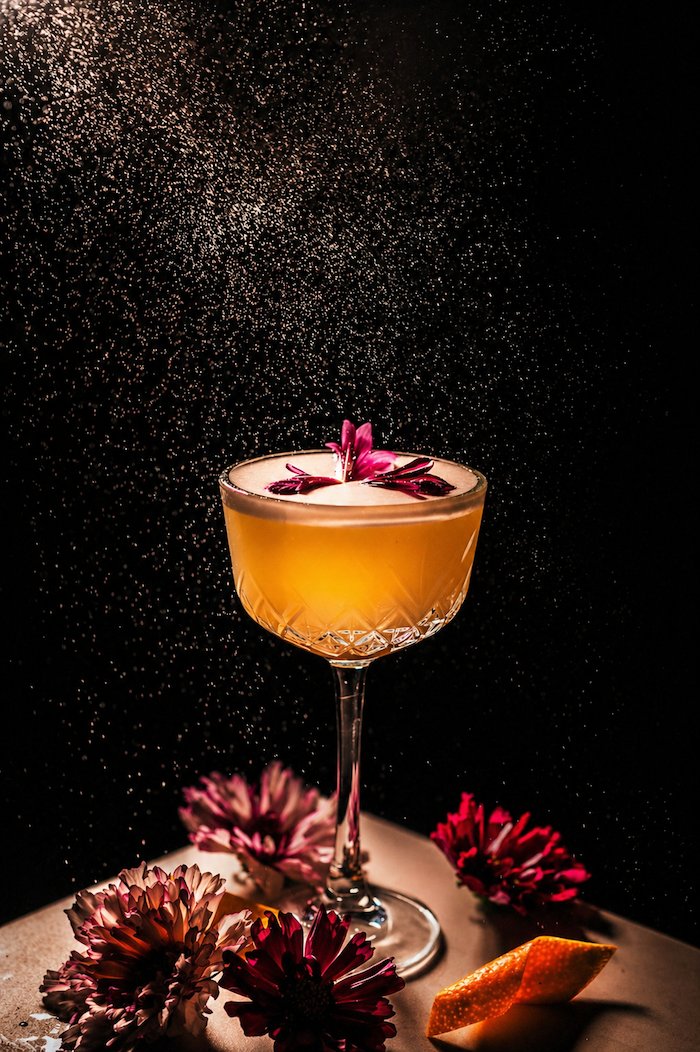 A coupe cocktail with edible flowers, lit from above with a fine mist of citrus oil.